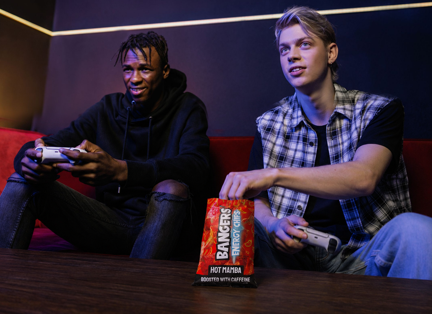 Two friends playing console games with a Hot Mamba Energy Chips bag between them on the coffee table as one reaches inside